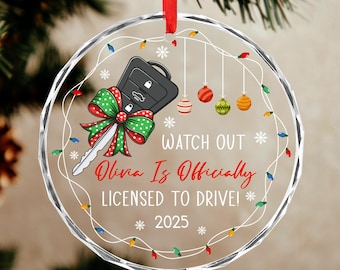 May include: Clear, round Christmas ornament with a car key design and a red ribbon. The key has a green and red bow. Text reads "Watch Out Olivia Is Officially Licensed to Drive! 2025". Decorative lights and ornaments surround the text.