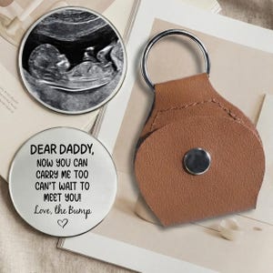 May include: A silver round keepsake with the text "DEAR DADDY, NOW YOU CAN CARRY ME TOO CAN'T WAIT TO MEET YOU! Love, the Bump" and a heart symbol. A brown leather keychain and a sonogram image are also in the image.