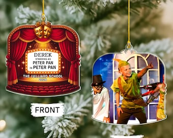 Puede incluir: Un adorno colgante de dos caras con un diseño de escenario de teatro en el frente, cortinas rojas, máscaras doradas y el texto DEREK STARRING AS PETER PAN IN PETER PAN THE JUILLIARD SCHOOL 2026. El reverso muestra una foto de una persona con disfraz verde.