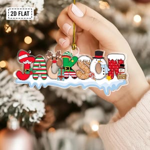 Puede incluir: Un adorno navide&ntilde;o plano de acr&iacute;lico transparente con el nombre JACKSON en letras coloridas con motivos festivos. Cada letra incorpora dise&ntilde;os como Santa, un reno, un elfo, un mu&ntilde;eco de nieve y regalos, con el a&ntilde;o 2025 en un letrero.