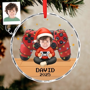 May include: A personalized Christmas ornament featuring a child in a Santa hat holding a game controller, set against a red game controller background adorned with Christmas lights. The ornament includes the name "DAVID" and the year "2025".