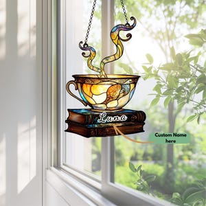 Op de afbeelding: Een glas-in-lood zonnevanger met een theekopjesontwerp met een handvat, rustend op twee boeken. De theekop heeft een kleurrijk glas-in-lood ontwerp met de naam "Luna" op de boeken. De zonnevanger hangt aan een ketting.