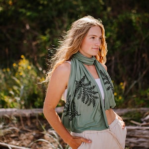 May include: Sage green scarf with a black fern leaf design. The scarf is draped around the neck and shoulders. The person is wearing a white top and tan pants. The background is blurred with green foliage.