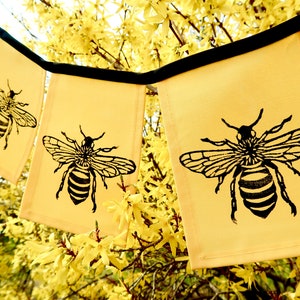 May include: A yellow fabric banner with black bee silhouettes printed on each flag. The banner is hanging from a tree branch with yellow flowers.