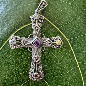 May include: A silver-toned pendant in the shape of a cross, adorned with small, round gemstones and a central square purple stone. The cross is set against a green leaf background, showcasing its intricate design and details.