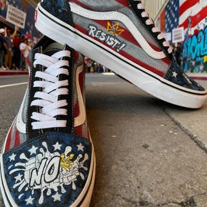 May include: Pair of custom Vans sneakers with a patriotic design. The shoes feature red and gray stripes, blue star accents, and the word "RESIST!" in white. The toe of the shoe has the word "NO" and a crown design.