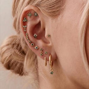Könnte beinhalten: Eine Kollektion von goldenen Ohrringen mit kleinen grünen Edelsteinen in verschiedenen Formen, darunter Sternmotive, Hänger und Stecker, die entlang des Ohrknorpels und des Ohrläppchens angeordnet sind und als Set getragen werden.