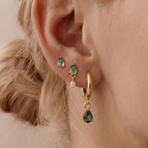 May include: A set of three gold earrings featuring green moss agate stones. The top earring is a small teardrop stud, the middle is a teardrop stud with a tiny clear crystal, and the bottom is a gold hoop with a dangling teardrop moss agate charm.