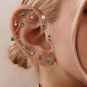 May include: A collection of gold-toned earrings featuring green moss agate stones in various shapes, including teardrop, marquise, and round cuts. The set includes studs, a chain-linked earring, and a dangling earring with three delicate gold chains.