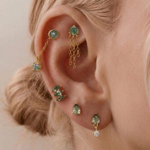 May include: A collection of gold-toned earrings featuring green moss agate stones. The set includes a chain-linked stud, a multi-chain dangle, a triple-stone cluster, a single pear-shaped stud, and a pear-shaped drop earring with a small clear stone.