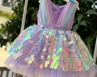 Op de afbeelding: Een lavendelkleurige jurk met iriserende pailletten en een tule rok. De mouwloze jurk heeft een V-hals en is versierd met pastelroze en blauwe bloemaccenten op de schouders. De jurk hangt aan een witte hanger.