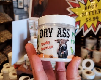 May include: A white jar with a black label that reads "DRY ASS" and a donkey illustration. The jar is labeled "body butter" and is being held by a hand. The background shows a sign with the text "THIS MOISTURIZER WILL HIDE THE FACT THAT YOU'VE BEEN TIRED SINCE 2010".