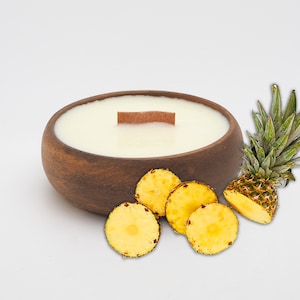 Op de afbeelding: Een houten kom kaars met een witte was bovenkant en een houten lont. De kaars is omringd door ananas plakjes op een witte achtergrond.