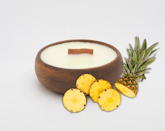 May include: A wooden bowl candle with a white wax top and a wooden wick. The candle is surrounded by pineapple slices on a white background.