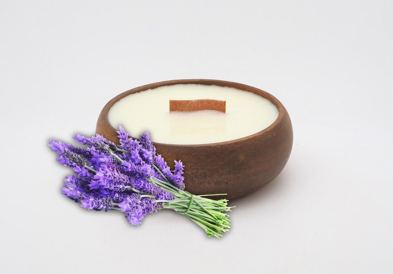 May include: A wooden bowl candle with a white wax top and a wooden wick. A bunch of lavender flowers are in the foreground.