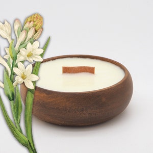 May include: A white candle with a wooden wick in a brown wooden bowl. The candle is surrounded by white flowers with green leaves.