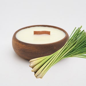 May include: A wooden bowl candle with a natural wood wick and a lemongrass scent. The candle is surrounded by a bunch of fresh lemongrass stalks.
