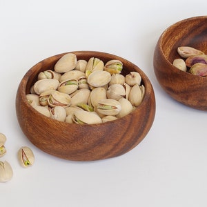 May include: Two wooden bowls, one filled with pistachios and the other with empty pistachio shells. The bowls are on a white surface.