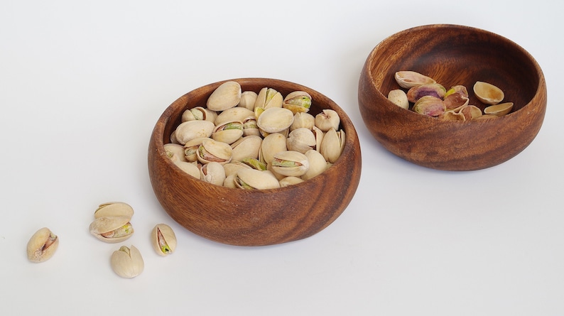 May include: Two wooden bowls, one filled with pistachios and the other with empty pistachio shells. The bowls are on a white surface.