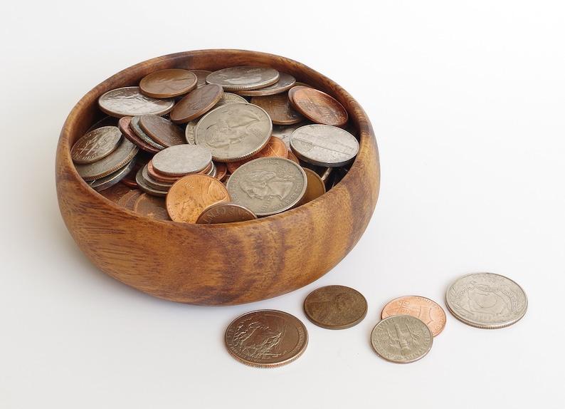 May include: A wooden bowl filled with a variety of US coins, including pennies, nickels, dimes, and quarters. The bowl is made of dark brown wood with a natural grain pattern.