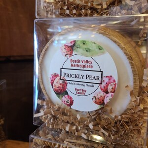May include: A white candle in a wooden container with a round label that says "Death Valley Marketplace" and "Prickly Pear" with a picture of a prickly pear cactus. The label also says "Made in Pahrump, Nevada" and "Pure Soy Candle".
