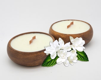 Puede incluir: Dos cuencos de madera llenos de velas blancas, cada uno con una mecha de madera. Los cuencos están rodeados de flores blancas de jazmín con hojas verdes.