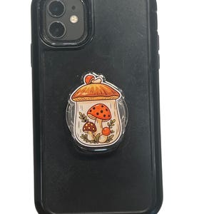 May include: A black smartphone case with a mushroom-themed pop socket. The pop socket features a cartoon illustration of a mushroom with a red and white spotted cap, surrounded by smaller mushrooms and leaves.