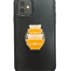 May include: Black phone case with a stack of yellow and white Pyrex bowls design. The bowls have a floral pattern. The phone case has a black textured surface and a camera lens cutout.