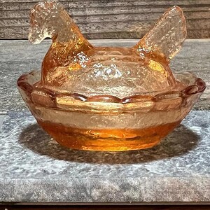 K&ouml;nnte beinhalten: Eine antike Glassch&uuml;ssel in Form eines Huhns mit einem gewellten Rand. Die Sch&uuml;ssel ist aus klarem Glas mit einer leichten Bernsteinf&auml;rbung. Das Huhn sitzt in einem Nest.