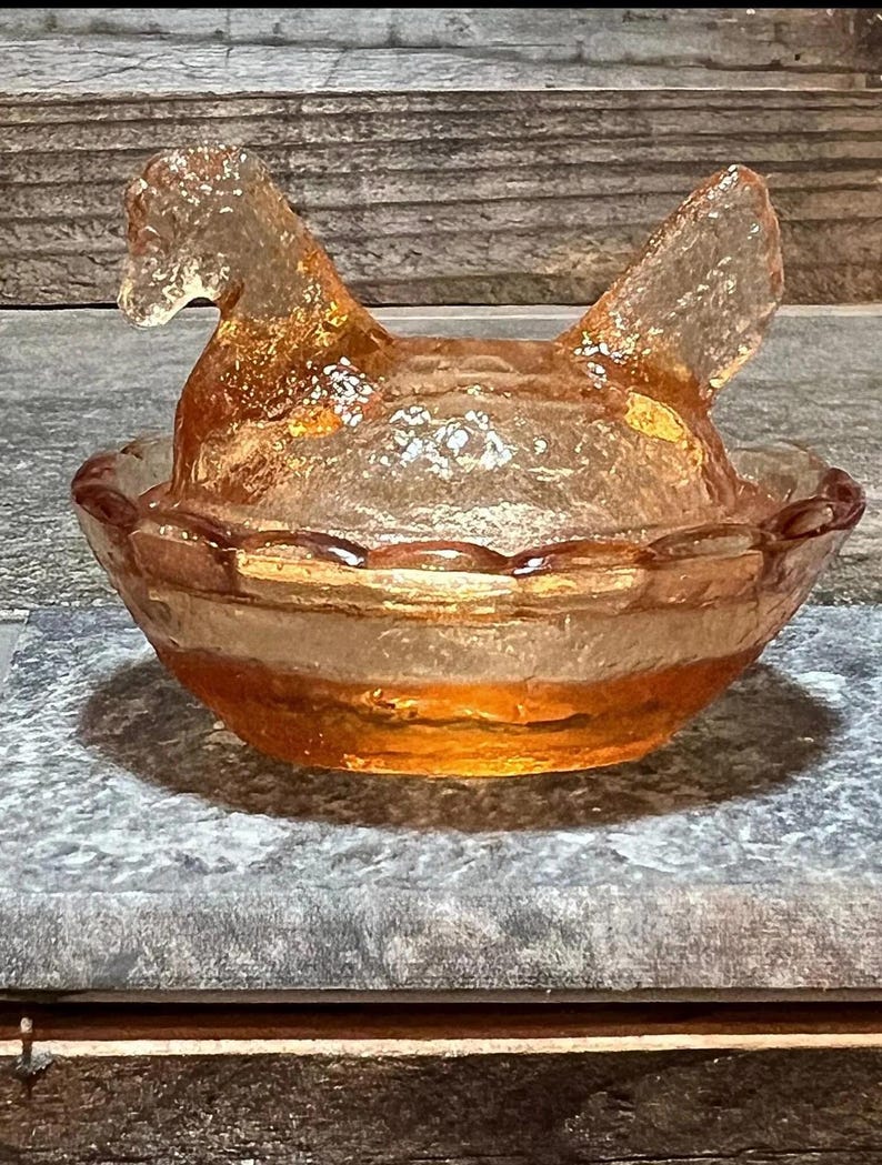 K&ouml;nnte beinhalten: Eine antike Glassch&uuml;ssel in Form eines Huhns mit einem gewellten Rand. Die Sch&uuml;ssel ist aus klarem Glas mit einer leichten Bernsteinf&auml;rbung. Das Huhn sitzt in einem Nest.