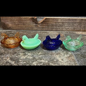 K&ouml;nnte beinhalten: Vier antike Nistschalen aus Glas in Form von H&uuml;hnern. Die Schalen sind in verschiedenen Farben, darunter Bernstein, Gr&uuml;n, Blau und Klar. Jede Schale hat ein geformtes H&uuml;hnerdesign.