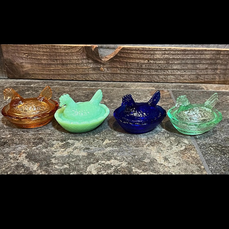 K&ouml;nnte beinhalten: Vier antike Nistschalen aus Glas in Form von H&uuml;hnern. Die Schalen sind in verschiedenen Farben, darunter Bernstein, Gr&uuml;n, Blau und Klar. Jede Schale hat ein geformtes H&uuml;hnerdesign.