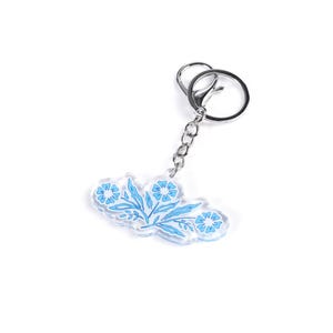 May include: A clear acrylic keychain featuring a blue floral design. The keychain has a silver-colored chain and clasp. The floral design includes flowers and leaves in shades of blue, creating a delicate and artistic aesthetic. The keychain is on a white background.