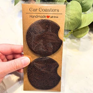 May include: Two brown leather car coasters with a paisley pattern. The coasters are packaged in a clear plastic bag with a brown card that says "Car Coasters Handmade with love".