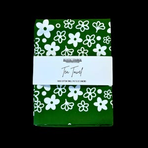 May include: A green tea towel with a white floral pattern. The towel is folded in half and has a label that reads "Rustic Timber Furniture & Home Tea Towel 100% Cotton Twill | 19.7 x 27.5 Inches".