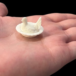 May include: A small, off-white figurine of a hen sitting in a decorative, scalloped dish. The hen has detailed feathers and a prominent tail. The figurine is held in a person's hand against a black background.