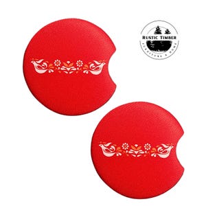 May include: Two red round beverage coasters with a white stylized design of birds and flowers. The coasters have a cut-out for a cup or glass. The text "RUSTIC TIMBER FURNITURE & HOME" is printed in black on a white circle.