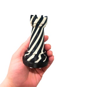May include: A black and white chess rook piece with a spiral design. The chess piece is held in a hand against a white background. The rook has a black base and a crenelated top.