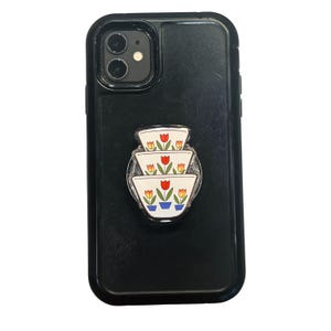 May include: A black phone case with a pop-up phone grip. The grip features a stack of three white bowls, each adorned with red, yellow, and orange tulips. The phone case is made of a textured material.