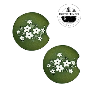 May include: Two green round coasters with a white floral design. The coasters have a cut-out for a cup or glass. The coasters are made by Rustic Timber Furniture & Home.