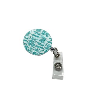May include: A round teal and white retractable badge reel with a decorative pattern. The badge reel has a clear plastic strap and a metal clip. The pattern includes stylized leaves, waves, and other shapes.