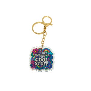 May include: A clear acrylic keychain with a gold-colored chain and clasp. The keychain features a colorful design with the text "IT'S NOT HOARDING IF IT'S COOL STUFF" surrounded by flowers, a rainbow, and peace signs.
