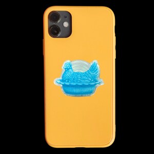 May include: A yellow phone case with a blue chicken and nest design. The chicken is in a nest-shaped holder, and the case has a glossy finish. The phone case is a protective accessory.
