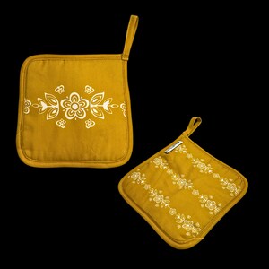 May include: Two mustard-yellow pot holders with white floral designs. One is square with a central floral pattern, and the other is diamond-shaped with diagonal floral stripes. Both have a loop for hanging.