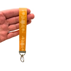 May include: Mustard yellow lanyard with a white floral pattern. The lanyard has a silver-colored metal clasp and a rectangular metal piece at the top. The lanyard is held in a hand against a white background.