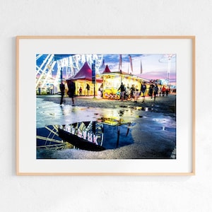 Puede incluir: Una vista nocturna de un carnaval con una noria, un puesto de comida iluminado y gente caminando. La noria se refleja en un charco en el suelo.