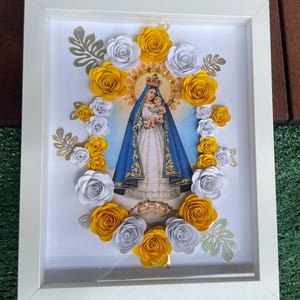 Puede incluir: Un marco blanco con una imagen de la Virgen María y el niño Jesús rodeada de flores de papel amarillas y blancas.