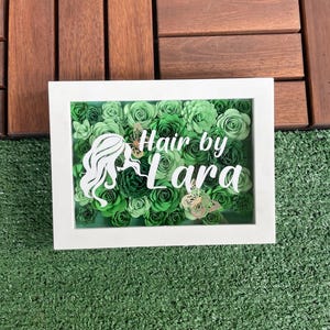 Puede incluir: Un marco blanco con un arreglo de flores de papel verde en su interior. Las flores están dispuestas en un patrón circular y hay una silueta de la cabeza y los hombros de una mujer en el centro. El texto "Hair by Lara" está escrito en letras blancas sobre las flores.