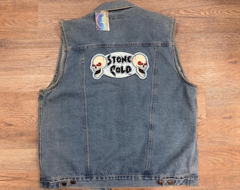 May include: A sleeveless denim jacket in a light blue wash. The back features a patch with two skull illustrations and the words "STONE COLD" in a light blue oval. A holographic tag is attached near the collar.