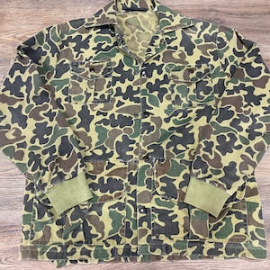 May include: A camouflage jacket in shades of green, brown, and black. The jacket features a button-up front, two chest pockets, and two lower pockets. The cuffs are a solid green color. The jacket is made of a durable fabric.